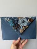 A slate blue fabric clutch bag with a triangular flap decorated with intricate beadwork, sequins, and embroidery in blue, gold, and brown, held by a hand against a plain light gray background.