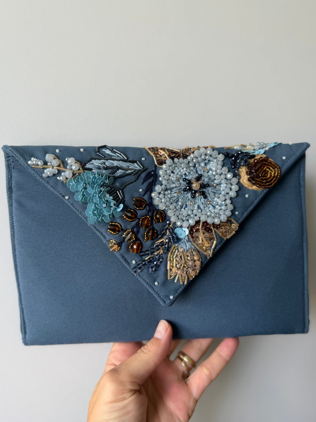 A slate blue fabric clutch bag with a triangular flap decorated with intricate beadwork, sequins, and embroidery in blue, gold, and brown, held by a hand against a plain light gray background.