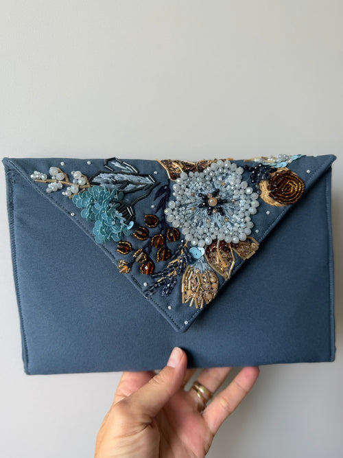 A slate blue fabric clutch bag with a triangular flap decorated with intricate beadwork, sequins, and embroidery in blue, gold, and brown, held by a hand against a plain light gray background.