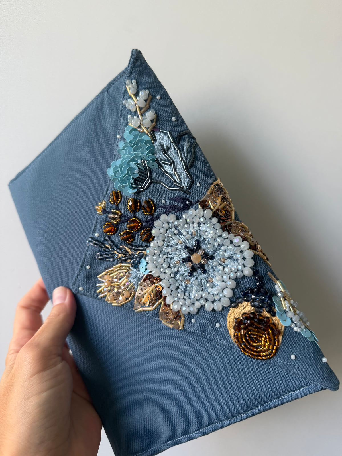 A slate blue fabric clutch bag with intricate floral embroidery, beadwork, and sequins in light blue, gold, and white, held by a hand against a plain light gray background.