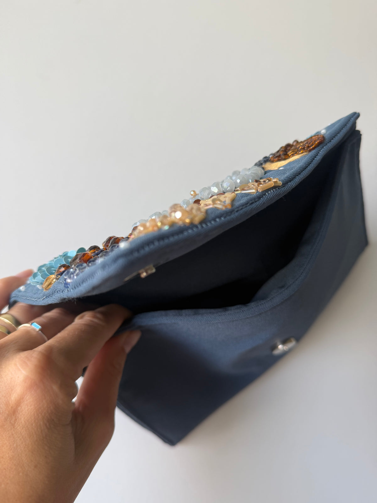 A navy blue fabric clutch bag with a hand holding the open flap, which is decorated with colorful rhinestone beadwork in shades of blue, gold, and brown.
